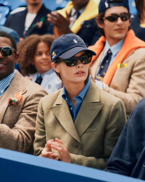 A woman in a crowd wearing a blazer, an Oxford shirt, and a baseball cap.