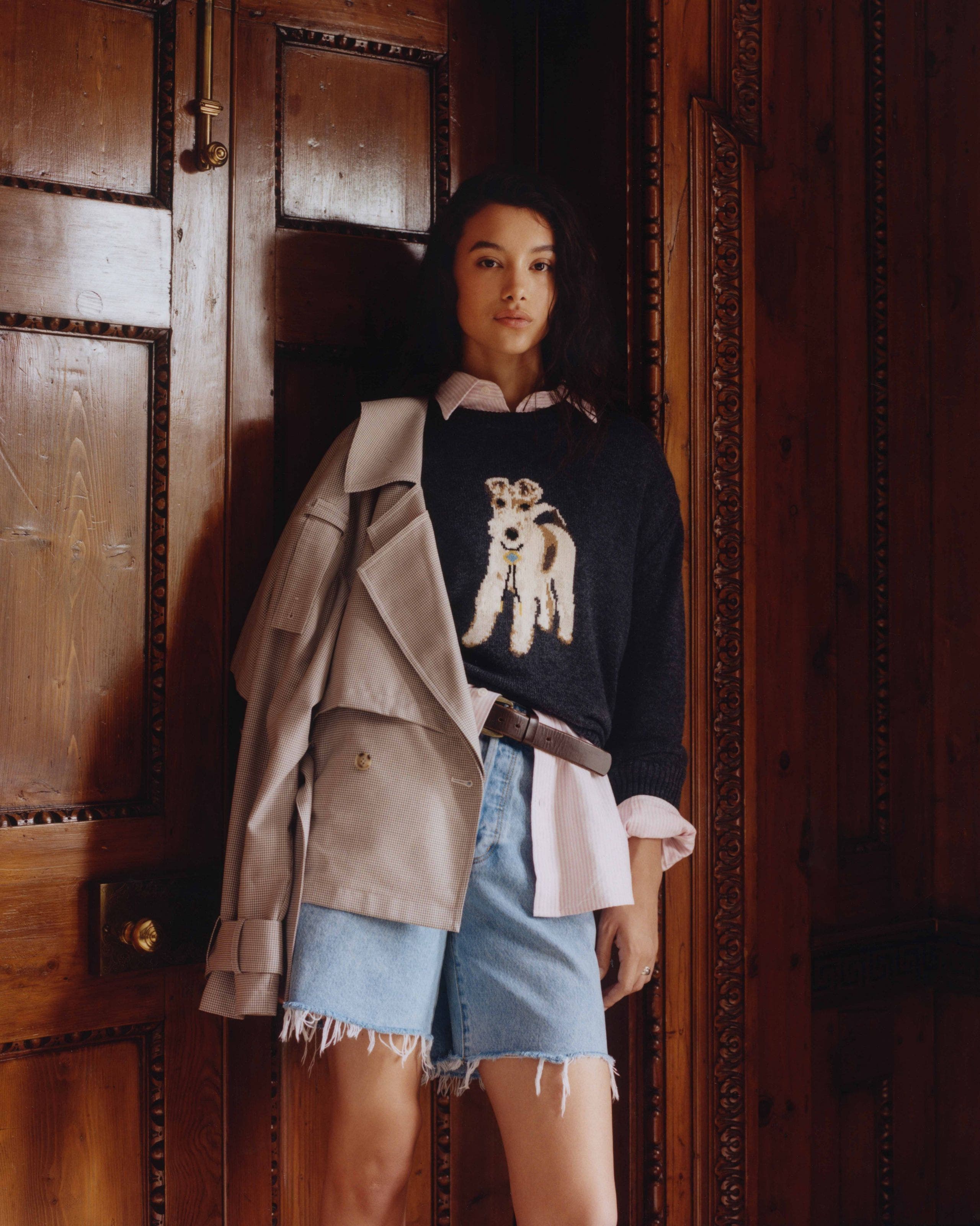 A woman wearing a sweater embroidered with a dog over a button up and cutoff denim jorts.