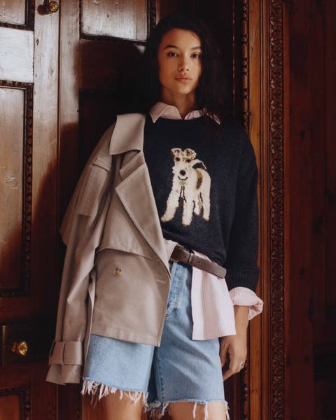 A woman wearing a sweater embroidered with a dog over a button up and cutoff denim jorts.