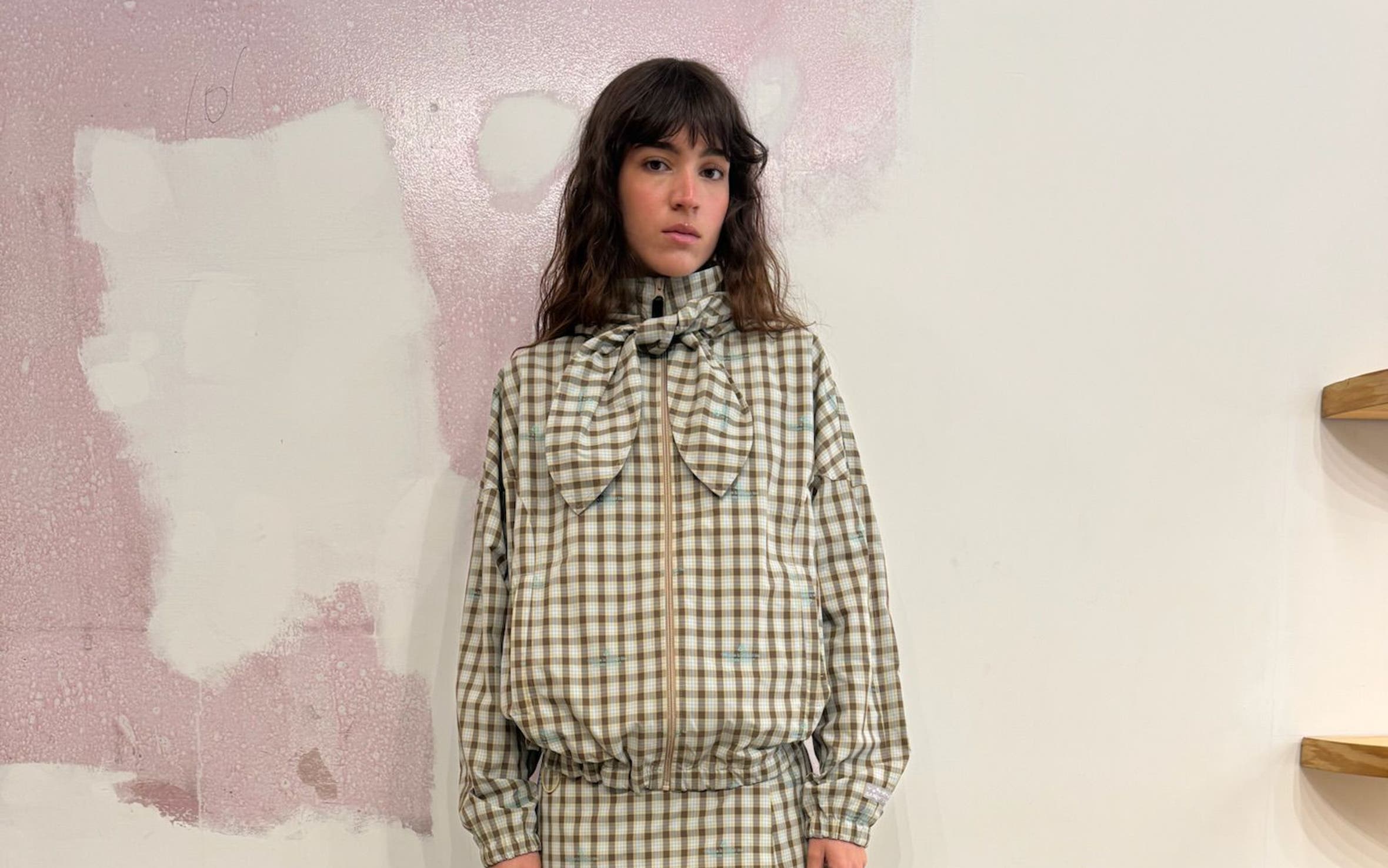 A model stands against a pink and beige textured wall, wearing a plaid button-up jacket in beige, blue, and cream tones with a high collar, chest pockets, and long sleeves, paired with a matching plaid mini skirt.