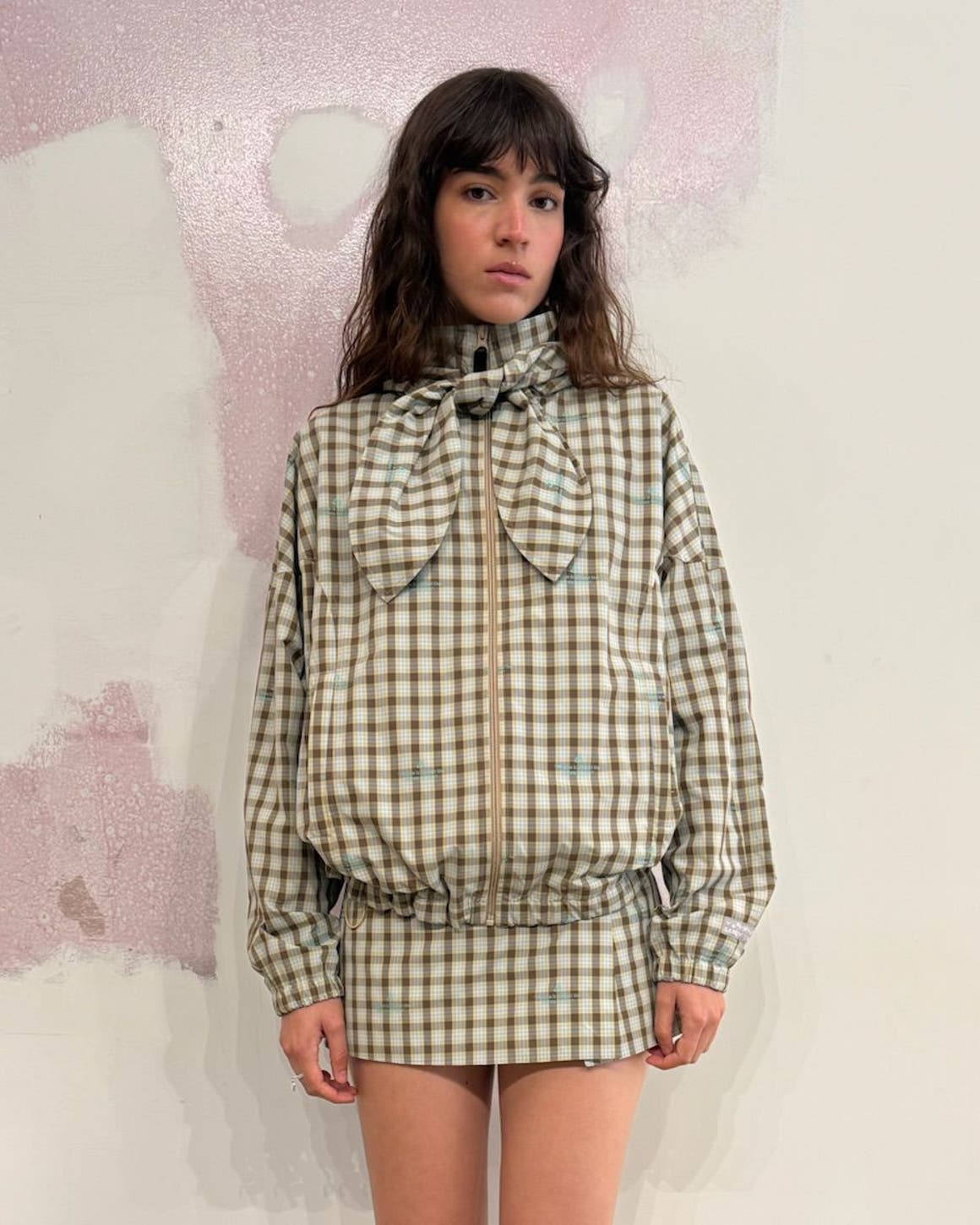 A model stands against a pink and beige textured wall, wearing a plaid button-up jacket in beige, blue, and cream tones with a high collar, chest pockets, and long sleeves, paired with a matching plaid mini skirt.