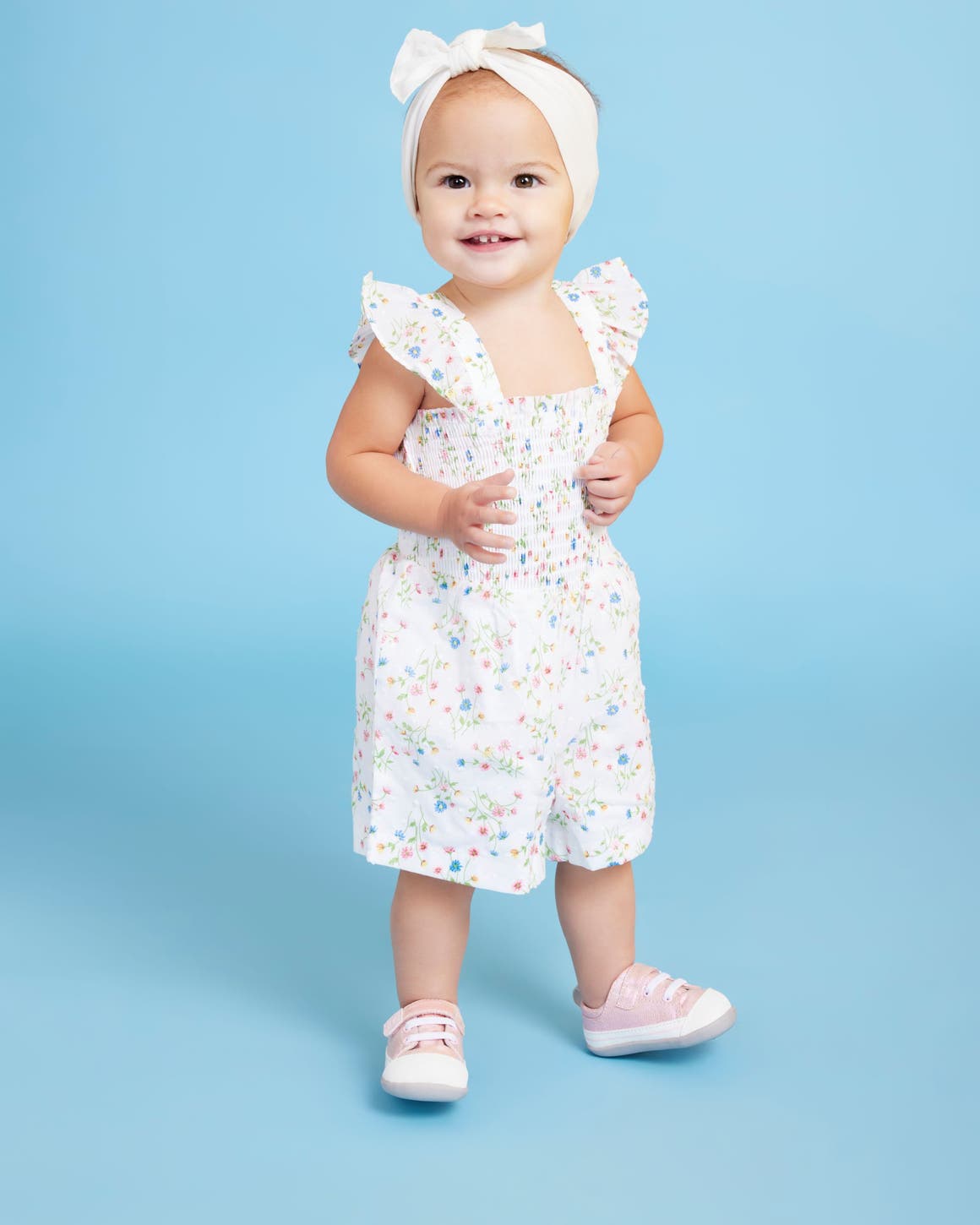 A toddler girl in pink sneakers and a floral romper.