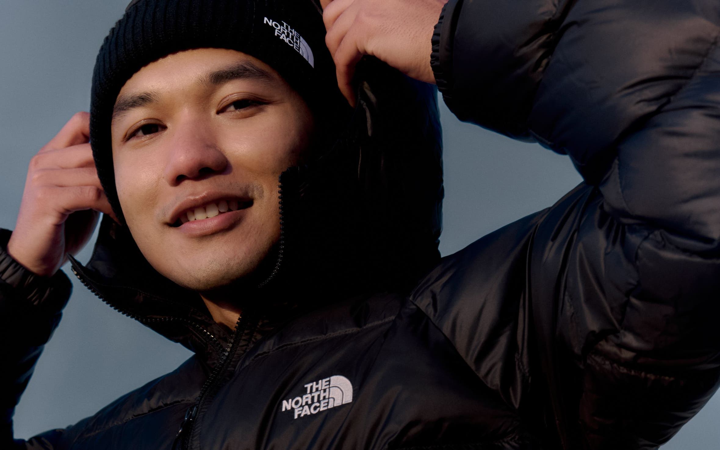 A man in a black The North Face puffer jacket and black knit beanie, pulling up the hood.