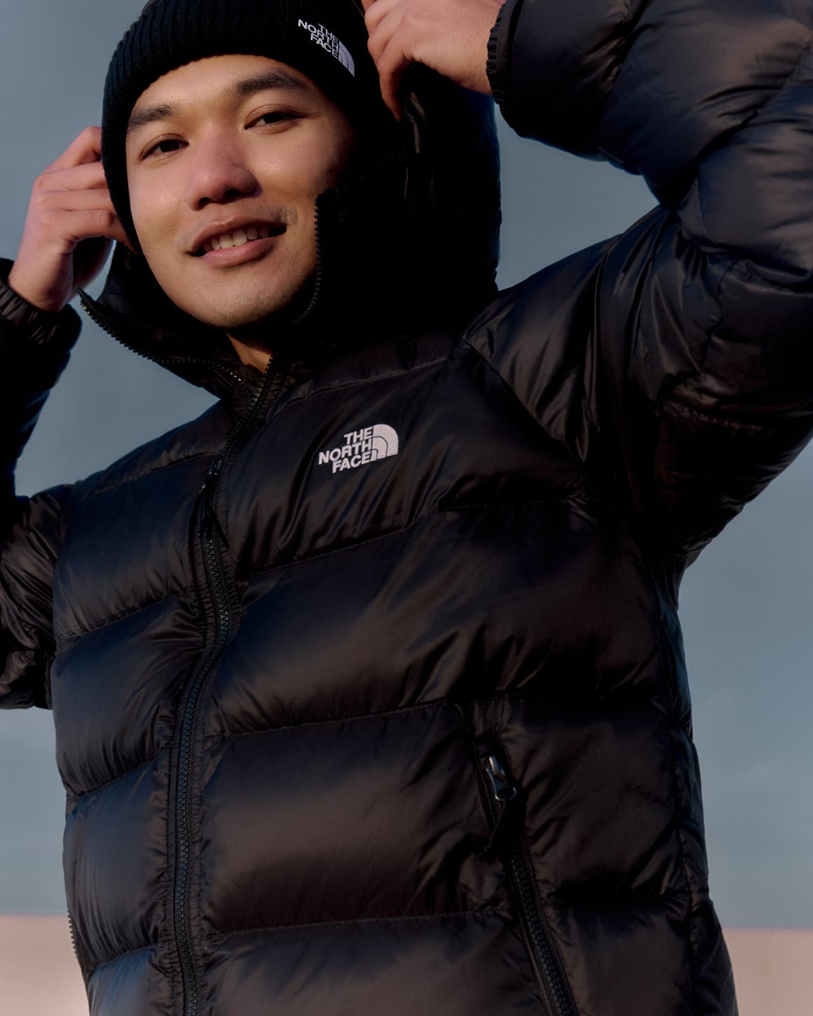 A man in a black The North Face puffer jacket and black knit beanie, pulling up the hood.