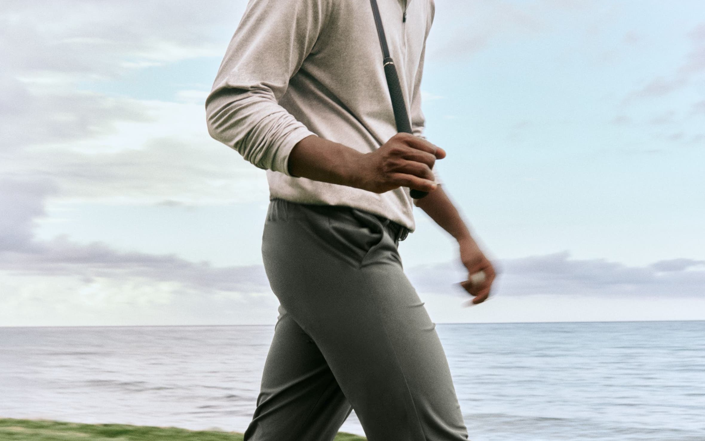 A man holding a golf club and wearing a quarter-zip sweater, polo shirt, trousers and sneakers.