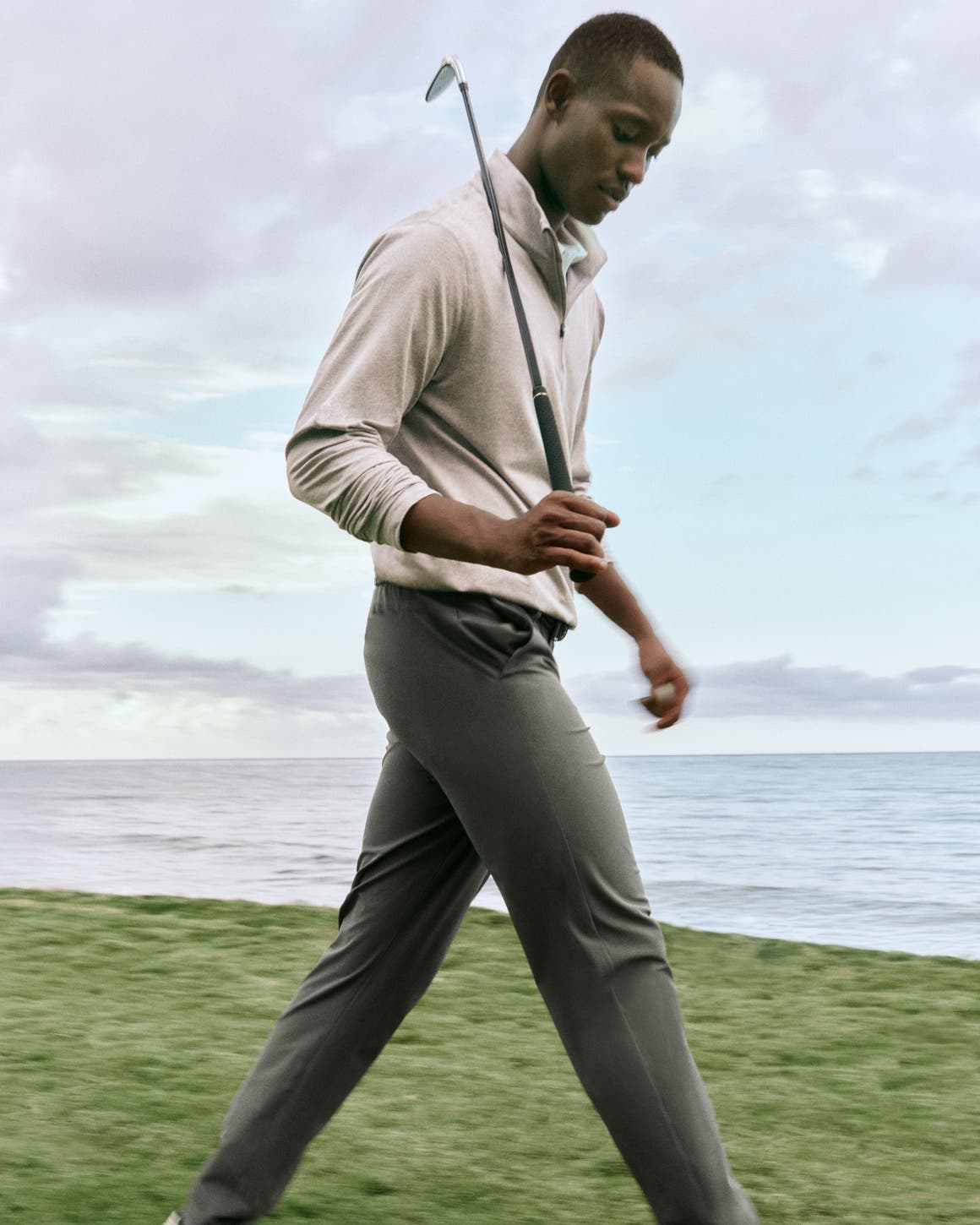 A man holding a golf club and wearing a quarter-zip sweater, polo shirt, trousers and sneakers.