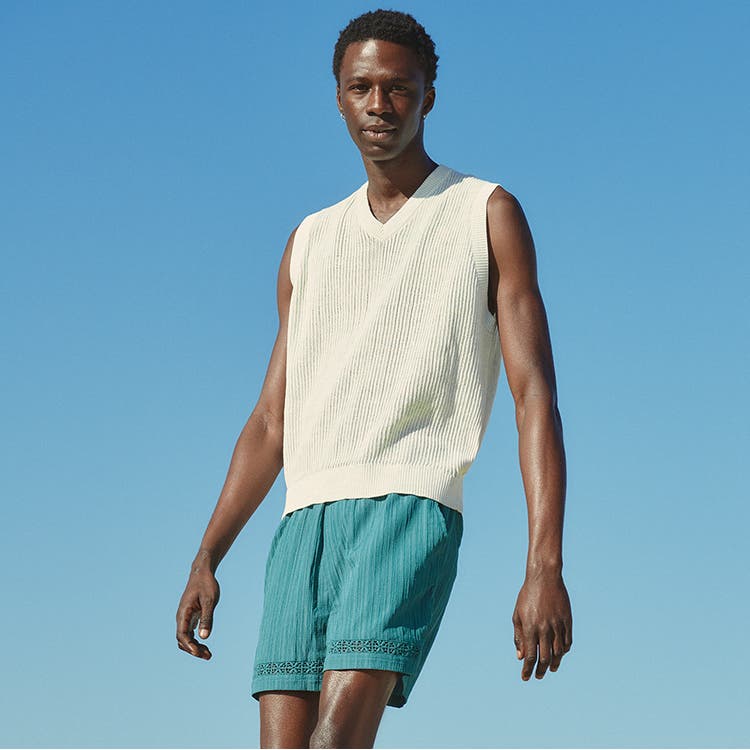 A model wearing a white sweater vest and turquoise shorts.