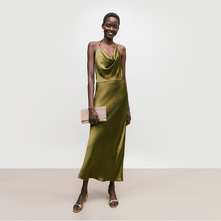 A woman wearing a wedding guest look: an olive slipdress, metallic sandals and a clutch.